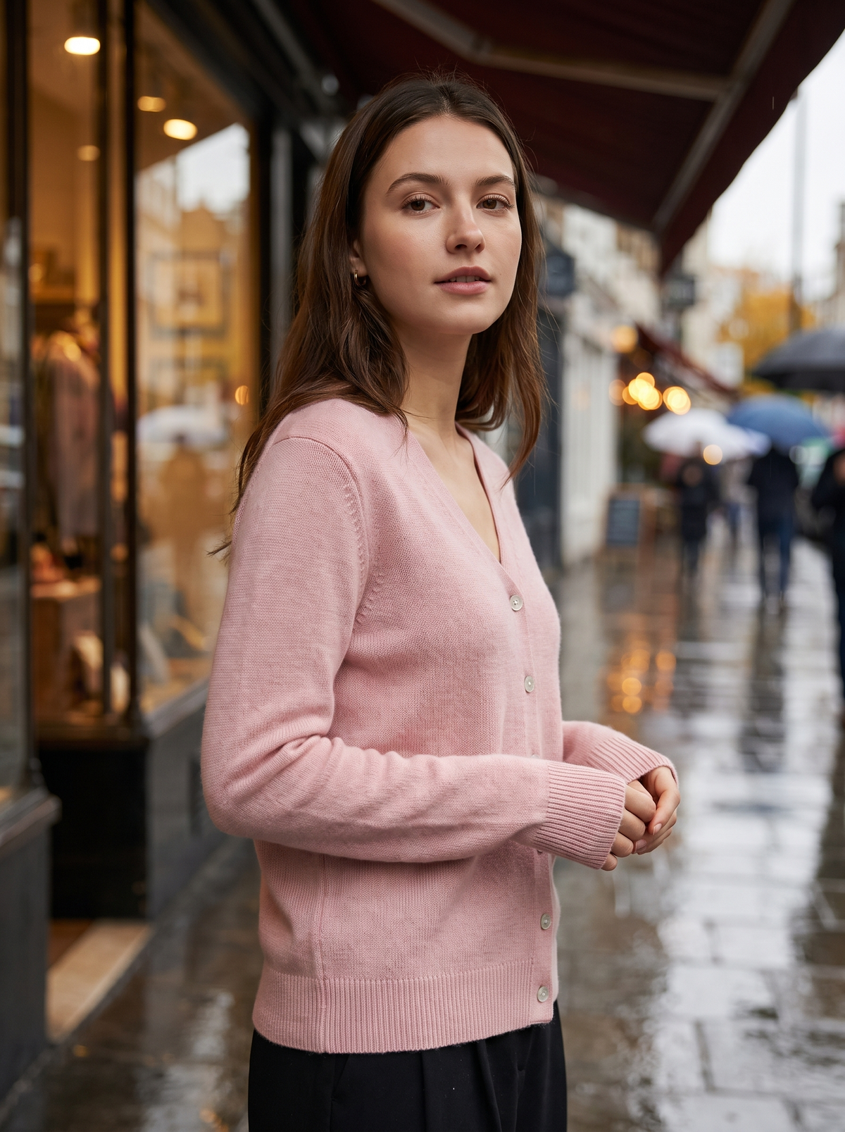 Henriette pink v neck cardigan worn by a model under a shop awning on a rainy street in autumn