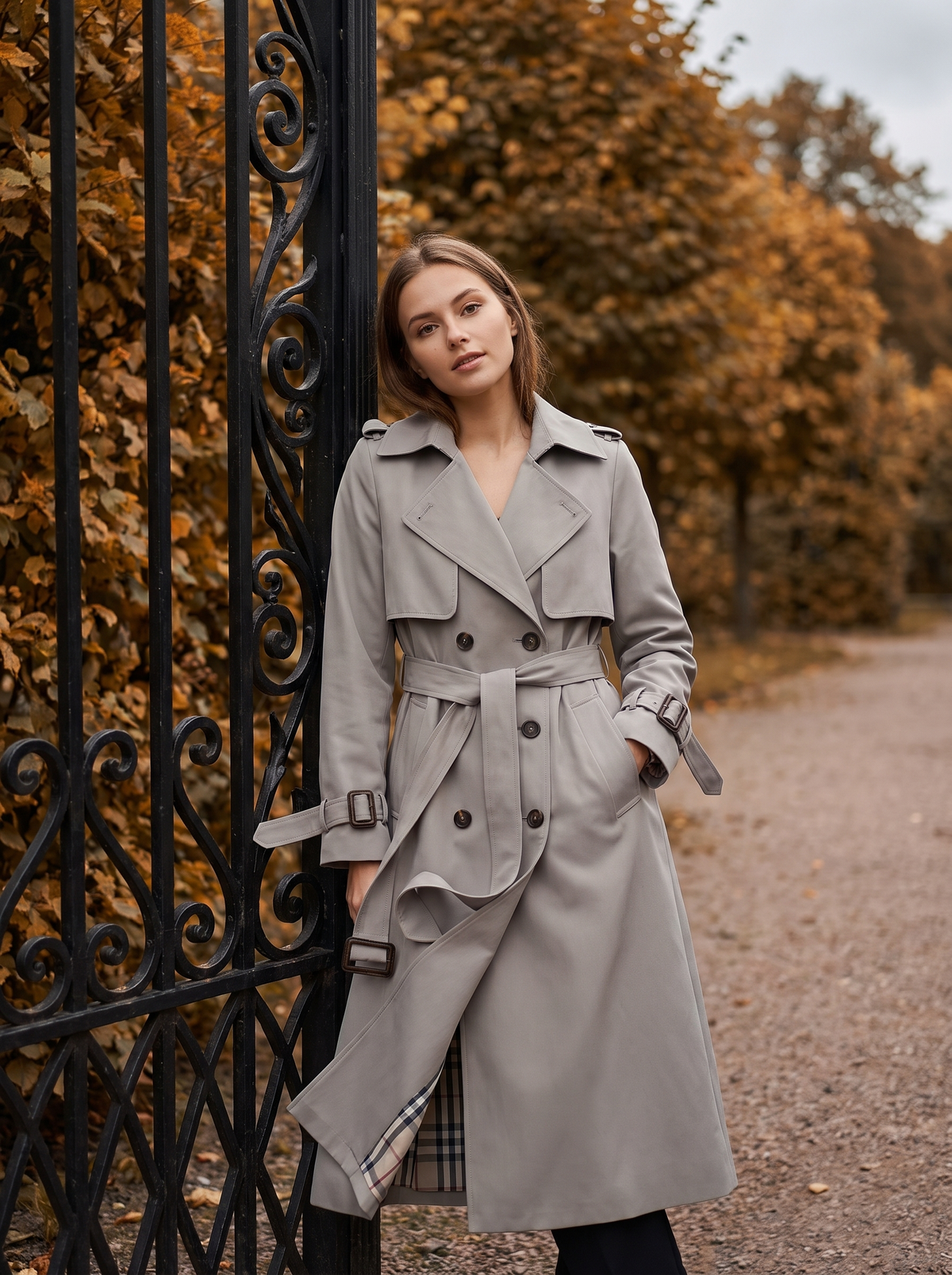 Aveline grey double breasted trench coat worn by a model at a park gate in autumn